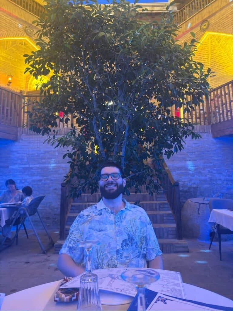 The author's partner, Ale, smiling at a restaurant table in Bukhara, Uzbekistan, framed by a tree and twinkling blue and yellow evening lights while waiting for dinner.