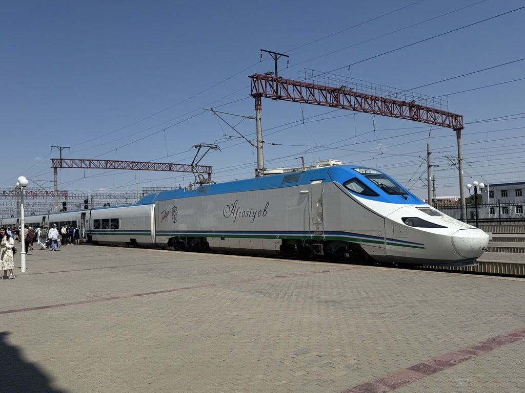 A wide-angle shot of the sleek white and blue Afrosiyob high-speed train parked along the platform at the Tashkent train station, with passengers walking alongside it preparing to board.