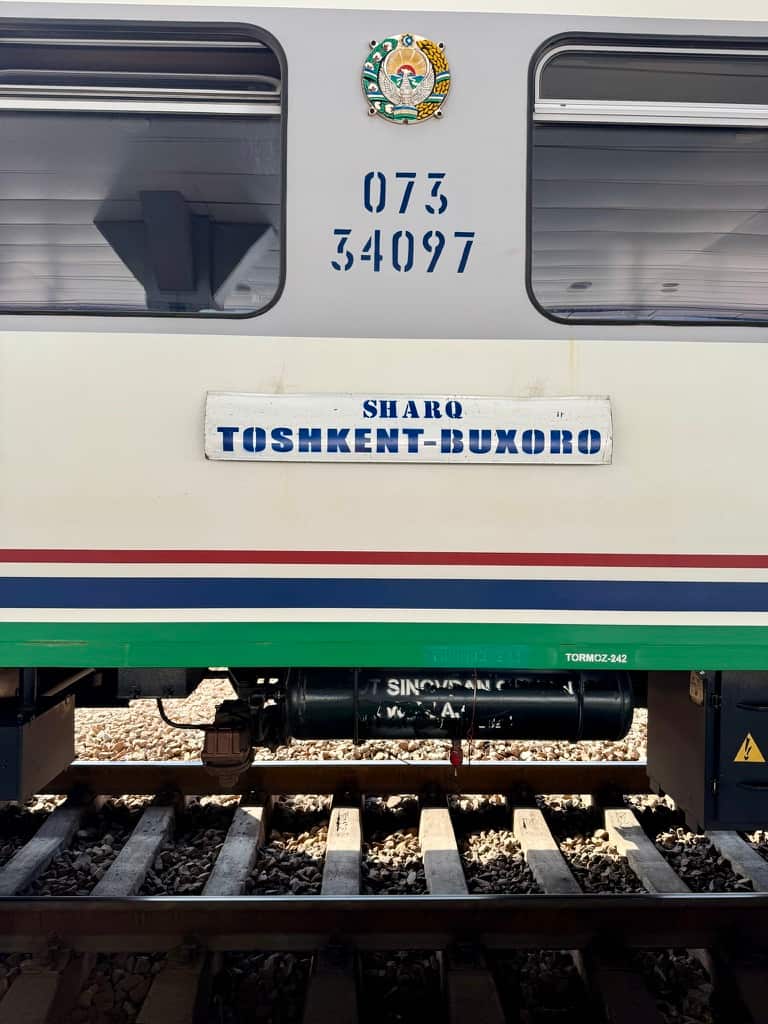 A closer exteriro shot of a Sharq express train car on the railway tracks, focusing on a rectangular route sign on the side that reads "SHARQ" with "TOSHKENT-BUXORO" printed directly below it.