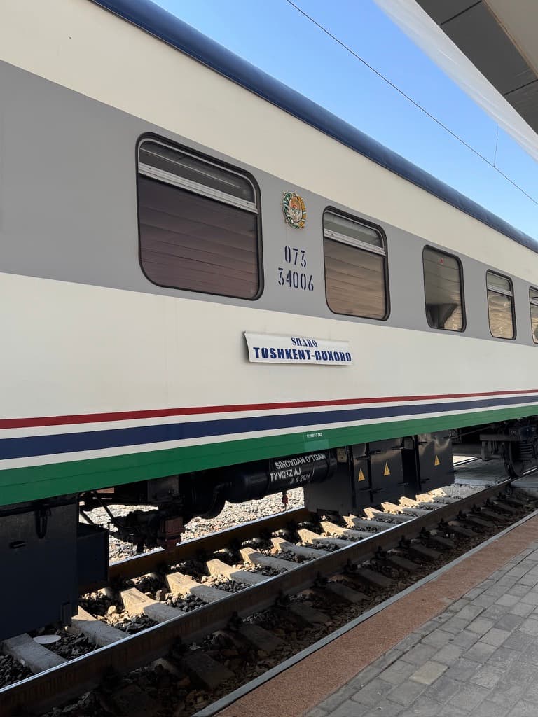An exterior profile shot of a Sharq express train car on the railway tracks, focusing on a rectangular route sign on the side that reads "SHARQ" with "TOSHKENT-BUXORO" printed directly below it.