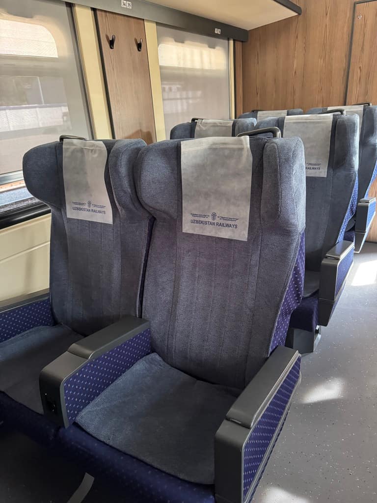 Interior view of the passenger coach on the Sharq express train in Uzbekistan, showcasing the seating arrangement with rows of fabric-covered chairs, wide windows, and an aisle walkway.