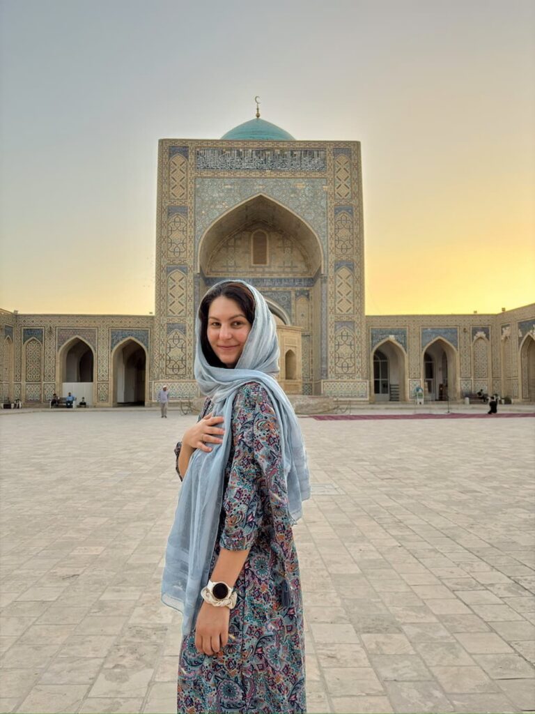 The author standing in front of a historic mosque in Bukhara, Uzbekistan, wearing a colorful dress and a blue hair covering that matches the intricate blue tilework and turquoise dome above.