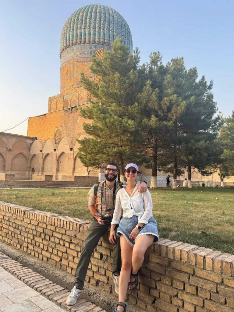 Two travelers standing in the stone courtyard of the Bibi-Khanym Mosque in Samarkand, Uzbekistan, with the massive turquoise blue dome and green trees visible in the background.