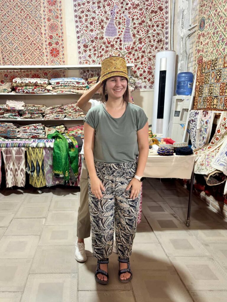Inside a colorful textile and souvenir shop in Bukhara, a local guide places a traditional Uzbek bridal cap decorated with intricate gold embroidery onto the author's head during a wedding attire demonstration.