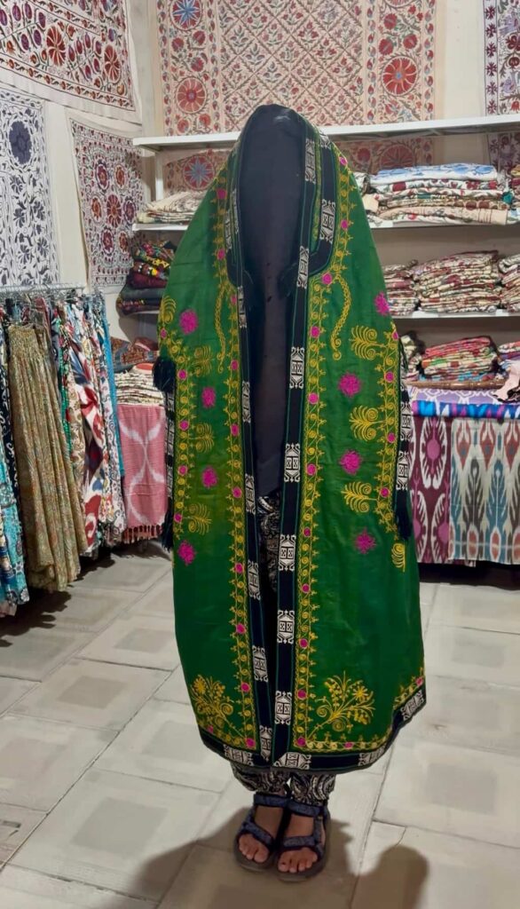 The author standing completely covered by a vibrant, beautifully patterned traditional Paranja robe draped over her head and entire body down to her feet, which remain visible, completing the historical Uzbek bridal attire demonstration.