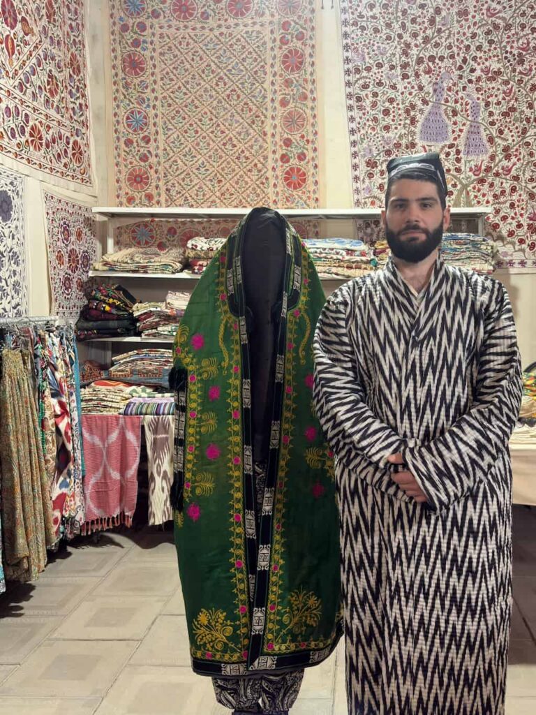 The author, fully dressed in a traditional historical Bukharian bridal robe and face veil, stands a step behind and to the right of Alessandro, who is wearing a magnificent, heavy Zarchopon robe richly embroidered with gold Zardozi threadwork and an iconic square Doʻppi cap with white geometric patterns.