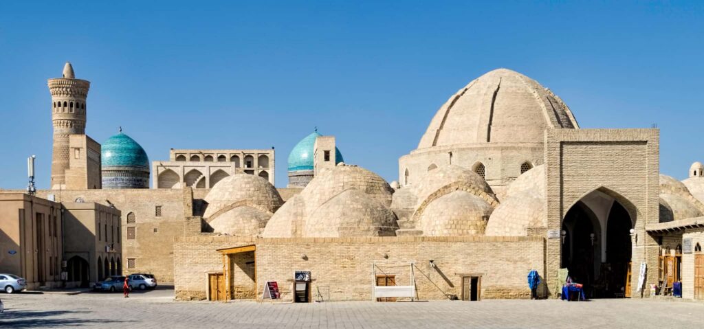 Panoramic landscape view of the earthen walls of the Ark of Bukhara, showing an arched entryway, historic brick domes, and a few local market goods on display.