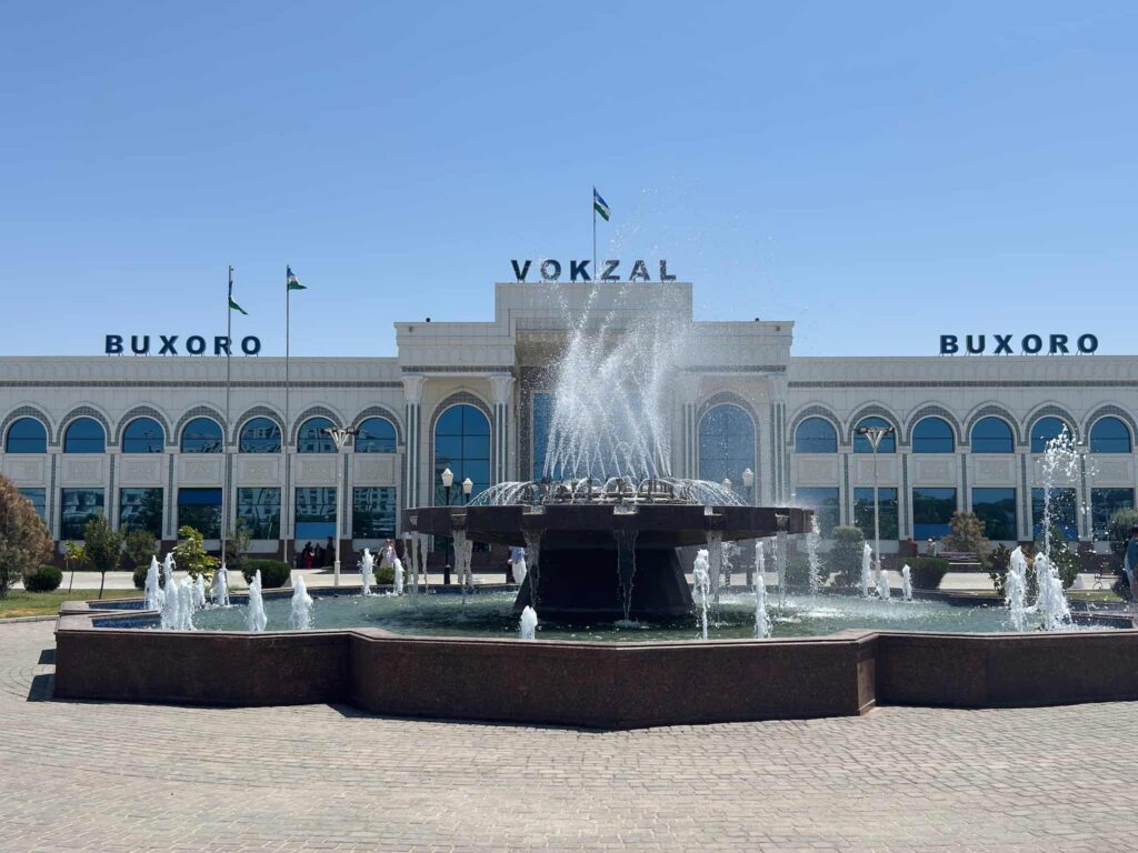 A centered landscape shot of the grand, white modern facade of the Bukhara railway station (Vokzal), with a large water fountain spraying in the square right in front of the main entrance.