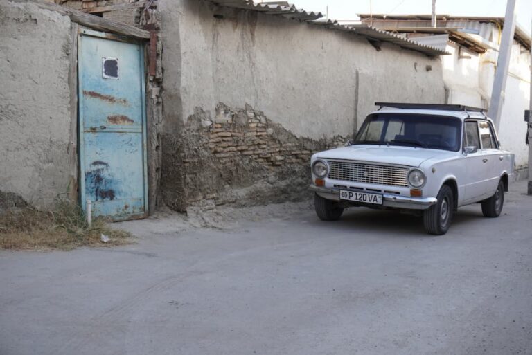 A narrow alleyway in Bukhara, Uzbekistan, featuring an old white car parked against an ancient brick wall next to a weathered blue wooden door.
