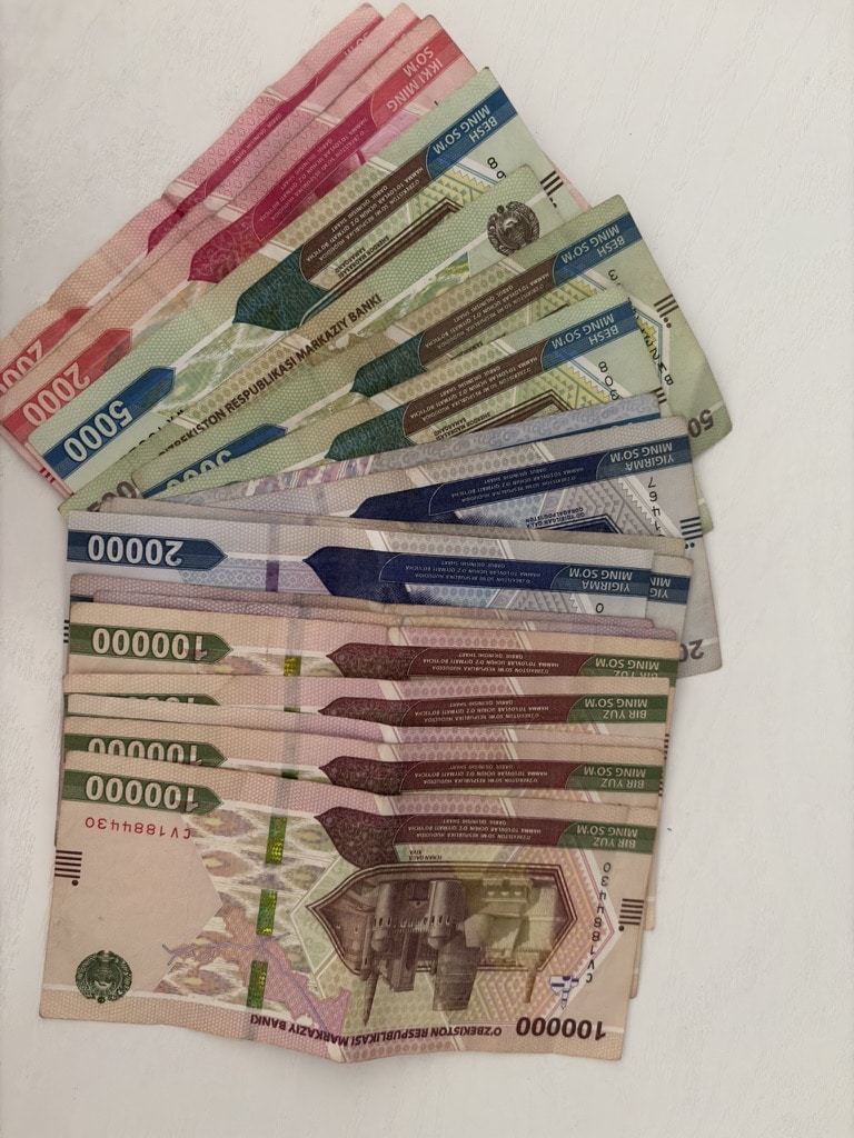 A top-down shot of several colorful Uzbekistani Soʻm banknotes arranged in a hand fan design on a clean white surface, showcasing the local paper currency.