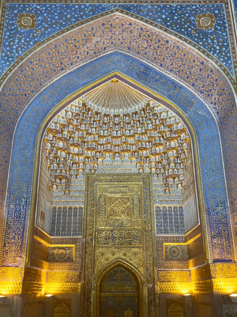 An interior view of a historic mausoleum gate in Samarkand, Uzbekistan, showcasing the grand portal archway with multiple stacked layers of intricate geometric designs in gold, brown, and deep blue.