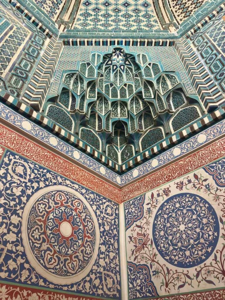 A split-view shot of an interior wall design inside a Shah-i-Zinda mausoleum, showcasing a vibrant green mosaic pattern on one half and a contrasting blue and red geometric tile design on the other, in Samarkand, Uzbekistan.