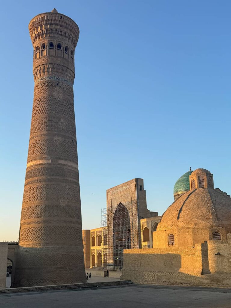 warm tones of the tall brick Kalan Minaret standing alongside the grand domes and facades of the Po-i-Kalyan complex against a brilliant golden sunset sky in Bukhara.