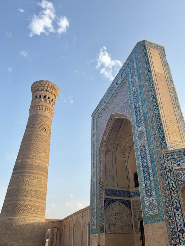 Low-angle view looking up at the grand tiled portal entry of the Po-i-Kalyan Mosque with the tall, intricate brown brick Kalan Minaret rising beside it against the bright blue sky.