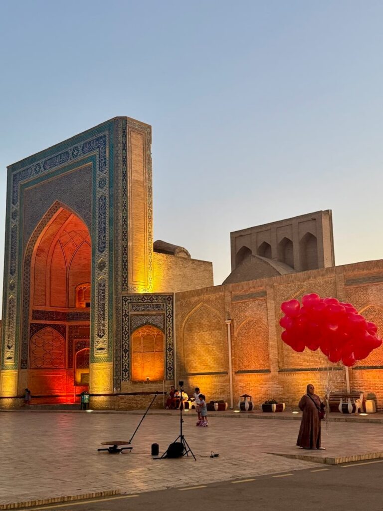 Side-angle view of the majestic Po-i-Kalyan Mosque illuminated at night, with a woman standing outside the complex holding bright red, heart-shaped balloons.