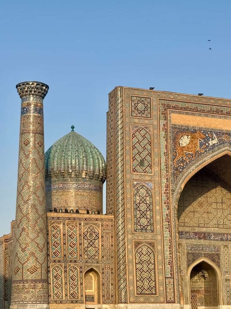 A close-up architectural shot of the historic Sher-Dor Madrasah in Registan Square, Samarkand, showing half of the grand arched entrance featuring its famous mosaic of a lion chasing a deer, flanked by a towering round minaret.