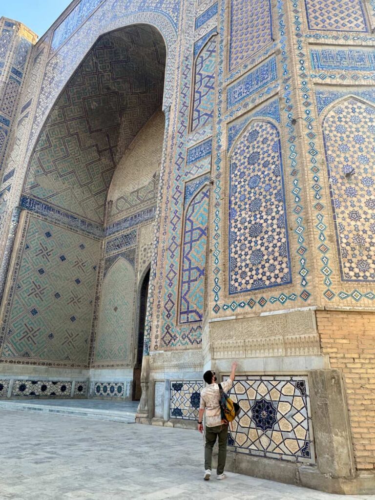 Close-up of a tourist placing their hand against the massive tiled wall of a madrasah in Registan Square, Samarkand, providing a clear sense of scale against the towering, intricate mosaic architecture.