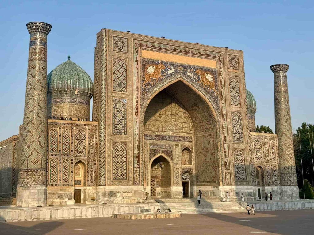 A side-angle view of the historic Sher-Dor Madrasah in Registan Square, highlighting the famous mosaic artwork of roaring lions under a rising sun for our Samarkand travel guide.
