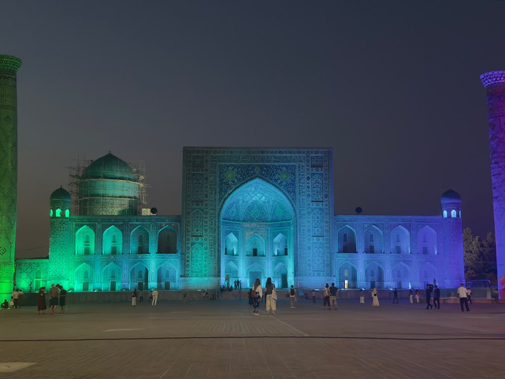 A focused night shot of a single madrasah facade at Registan Square in Samarkand, Uzbekistan, dynamically illuminated with vibrant pink, green, and blue modern lights right before an evening light show.
