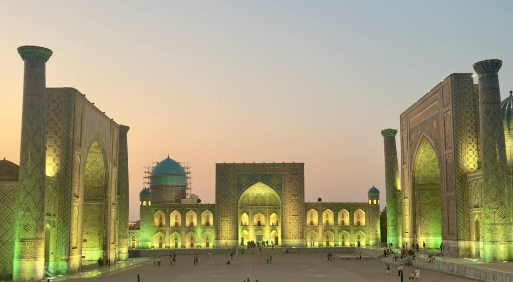A panoramic night view of the entire Registan Square in Samarkand, Uzbekistan, beautifully illuminated with warm yellow lights after sunset, showcasing all three historic madrasahs glowing against the dark twilight sky as featured in our Uzbekistan travel itinerary guide.