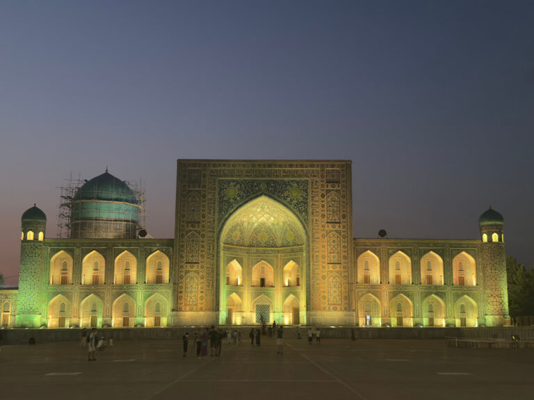 A focused night view of a magnificent madrasah facade at Registan Square in Samarkand, Uzbekistan, glowing under warm yellow illumination against a deep twilight sky after sunset.