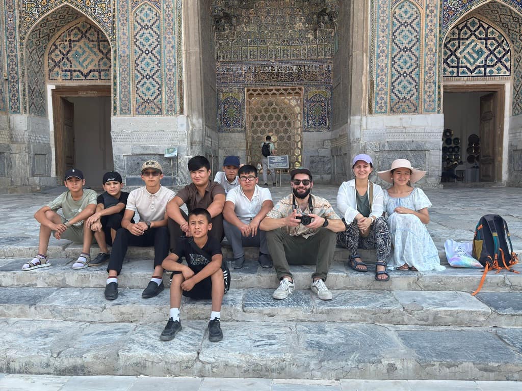 A group of young Uzbek children sitting with tourists and a tour guide in front of a historic madrasah at Registan Square, featured in this Samarkand travel guide.