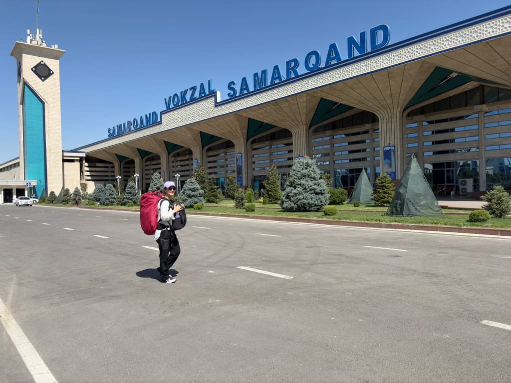 A traveler standing outside the modern facade of the Samarkand train station carrying two large travel backpacks, preparing for a train journey in Uzbekistan.