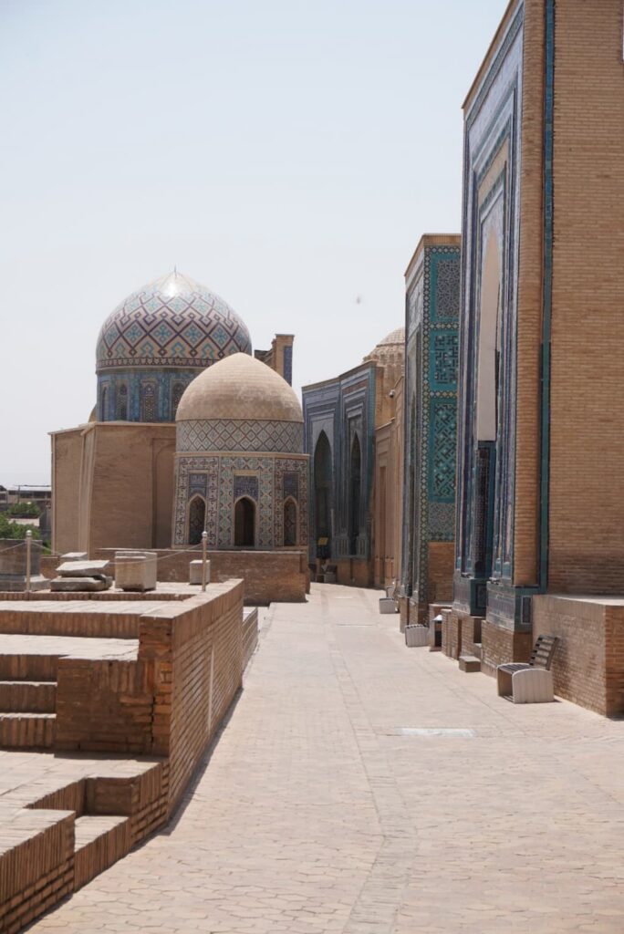 A scenic view looking down the stone pathway of the Shah-i-Zinda Necropolis in Samarkand, Uzbekistan, lined with a row of historic blue-tiled tombs and multiple prominent turquoise domes.