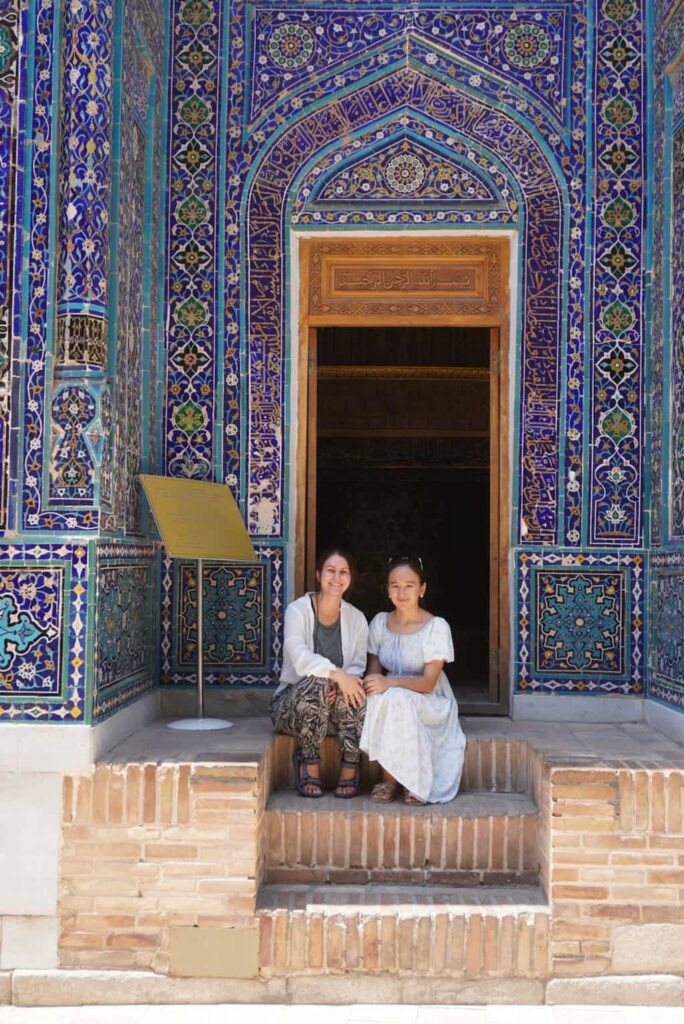 The travel blogger and a young local tour guide sitting together on the stairs in front of an intricately tiled blue mausoleum at the Shah-i-Zinda Necropolis in Samarkand, Uzbekistan.