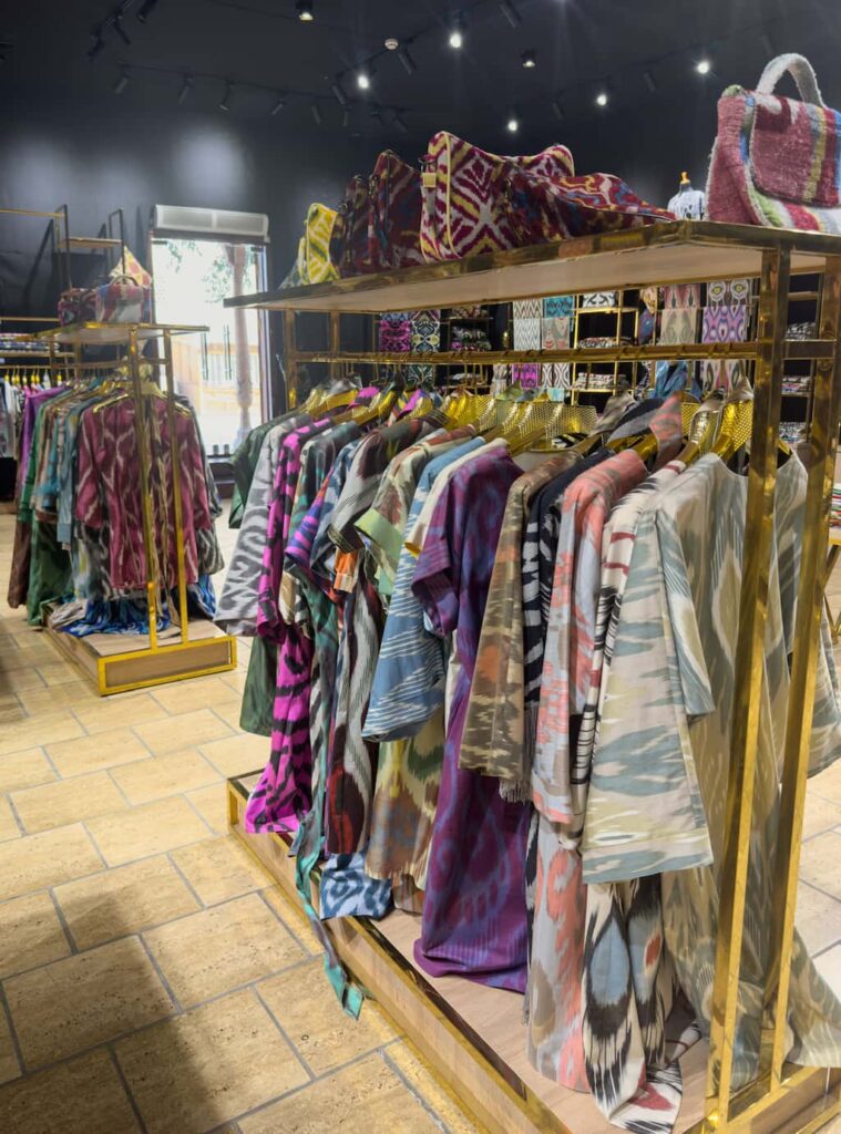 A vibrant display of colorful silk dresses and traditional patterned clothing hanging inside a local boutique fashion store in Bukhara, Uzbekistan.