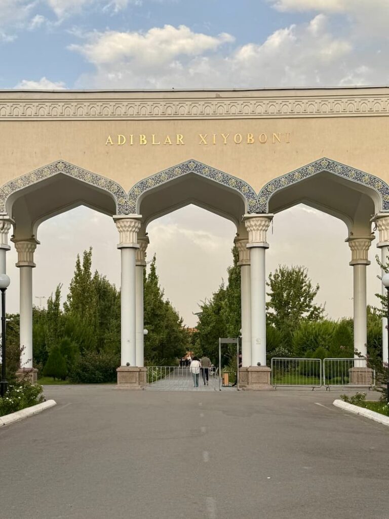 The grand entry to Magic City Park in Tashkent, featuring tall pillars and arches with a wide road leading through it and dense trees in the background
