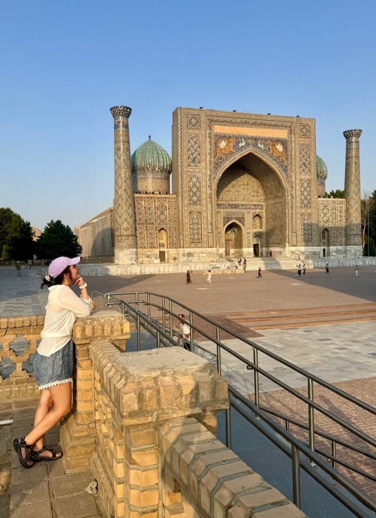 Rear view of the travel blogger overlooking the grand Sher-Dor Madrasah from an elevated viewpoint in Registan Square, Samarkand, capturing the iconic tiger mosaic facade from a unique side angle.