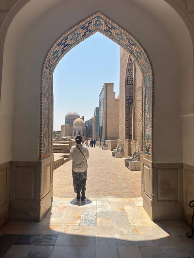 A traveler standing under a majestic arched portal, overlooking the stunning complex of blue-tiled tombs and turquoise domes at the Shah-i-Zinda Necropolis in Samarkand, Uzbekistan.