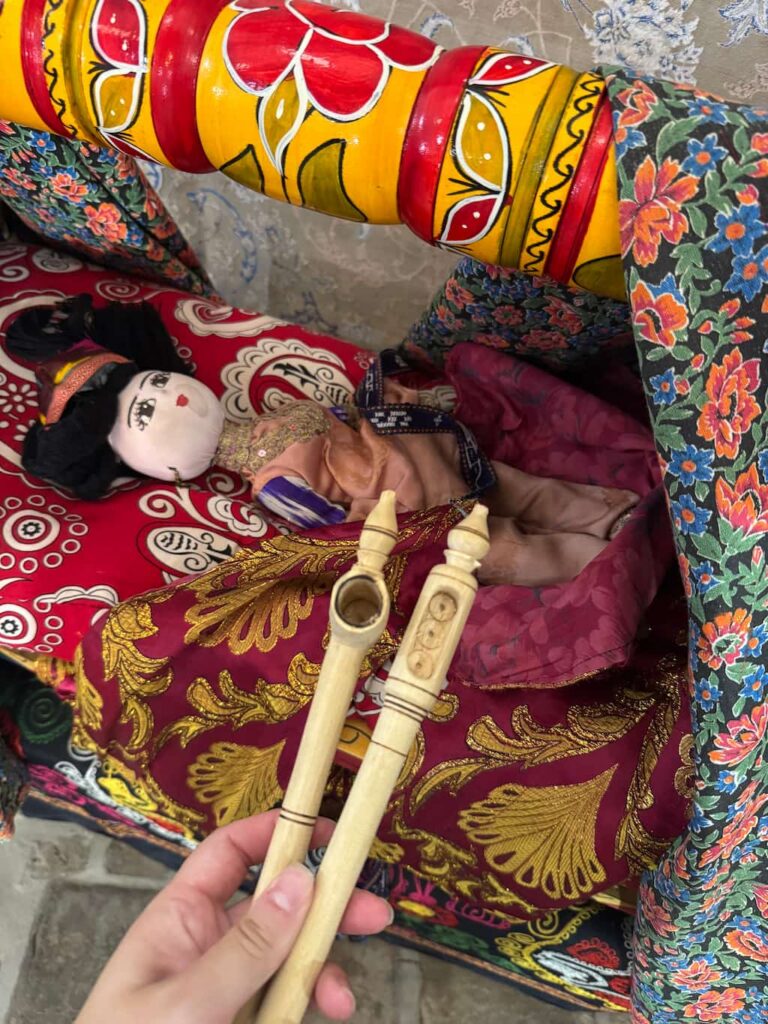 A close-up view of a traditional Uzbek wooden beshik cradle with a doll wrapped in colorful blankets, with the author holding two sumak tubes—one designed for boys and one for girls—used for directing urine out of the cradle.