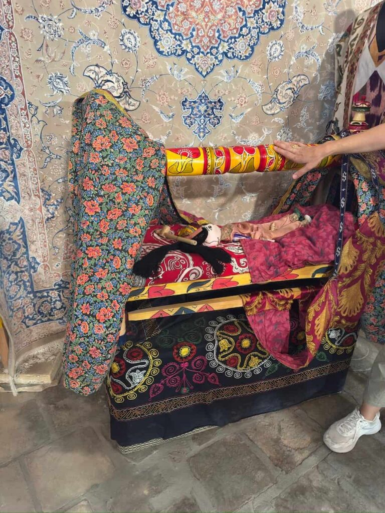 A tour guide in Bukhara demonstrating the use of a traditional Uzbek wooden cradle, known as a beshik, featuring a doll wrapped snugly inside under vibrant, colorful patterned blankets.