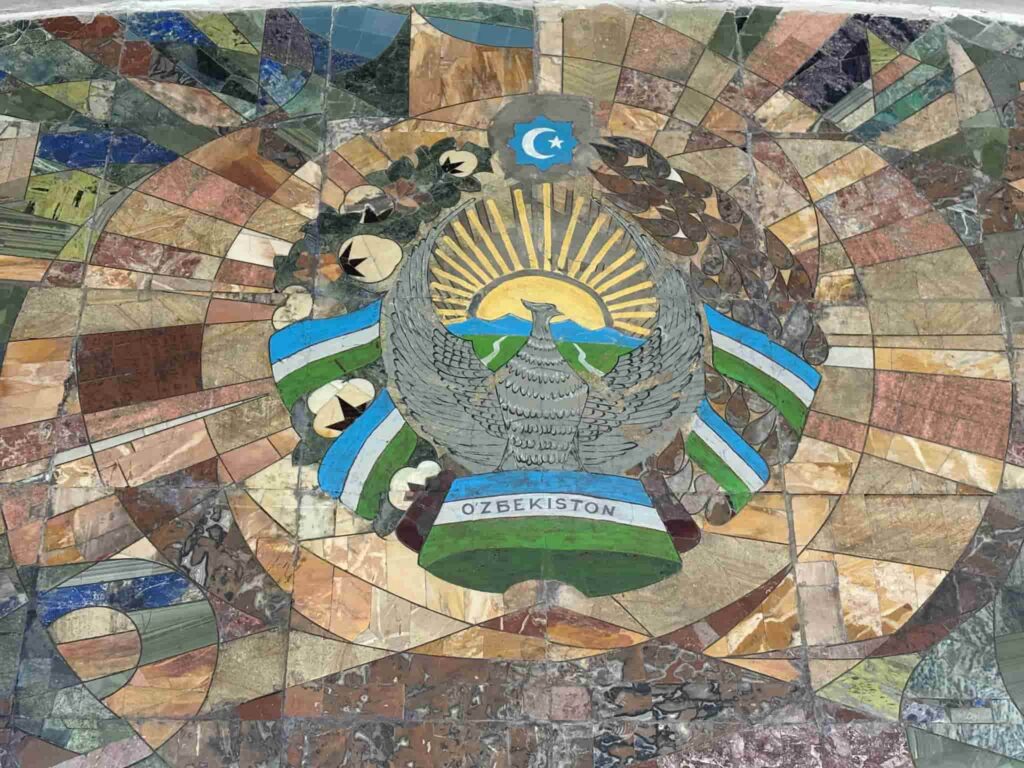 A mosaic mural at the entrance of Kosmonavtlar or Uzbekiston metro station in Tashkent, featuring the majestic Huma bird with outstretched wings over the vintage State Emblem of the Uzbek Soviet Socialist Republic, surrounded by woven cotton plants, wheat stalks, and a rising sun in a Soviet propaganda art style.