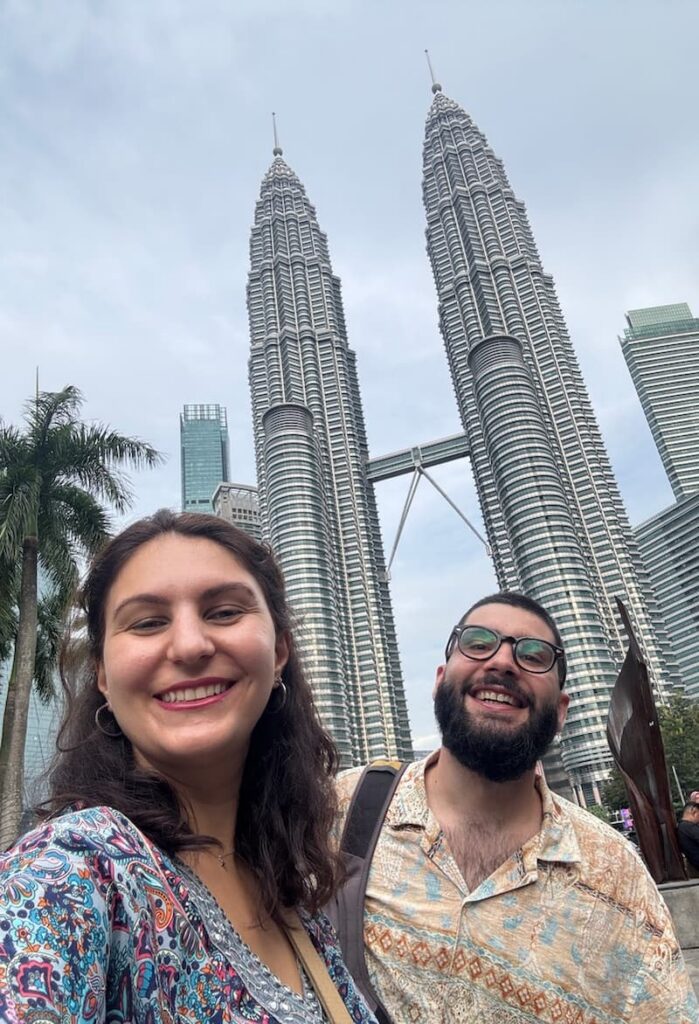 The authors of this travel blog smiling and taking a selfie from a low angle with the popular petronas towers in Kuala Lumpur, Malaysia
