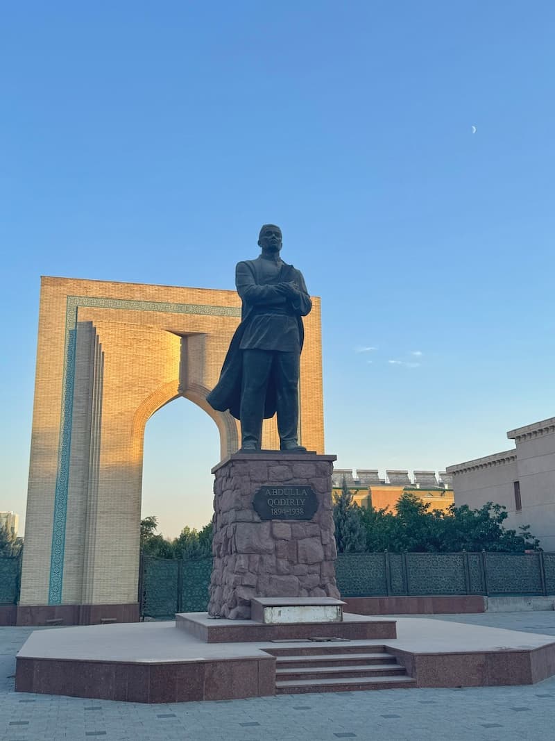 A bronze statue of Amir Temur standing before a massive, historic brown brick archway entrance, with the ancient structure sharply split between golden hour sunlight and deep shade under a clear blue sky.
