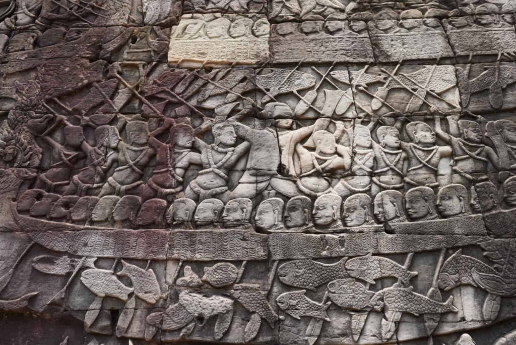 Intricate stone bas-relief carvings on a temple wall at Angkor, depicting ancient figures in a boat surrounded by fish.