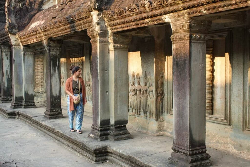 A traveler walking along a colonnaded corridor featuring ancient stone carvings at Angkor Wat.