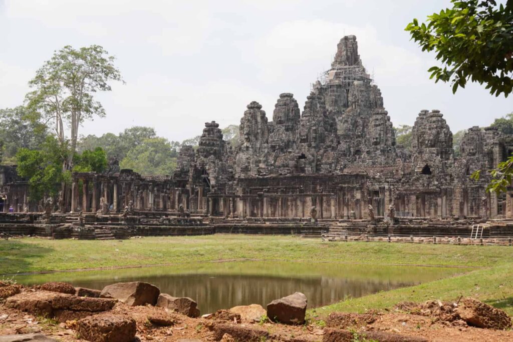 A grand view of the Bayon temple with its iconic stone faces rising behind a grassy foreground pool.