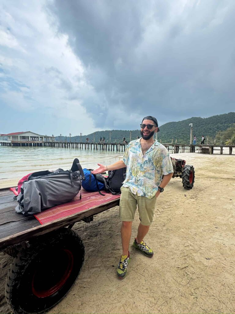 Ale, one of the blog's authors, smiling next to travel luggage piled on a beach cart with a wooden pier and ocean in the background.