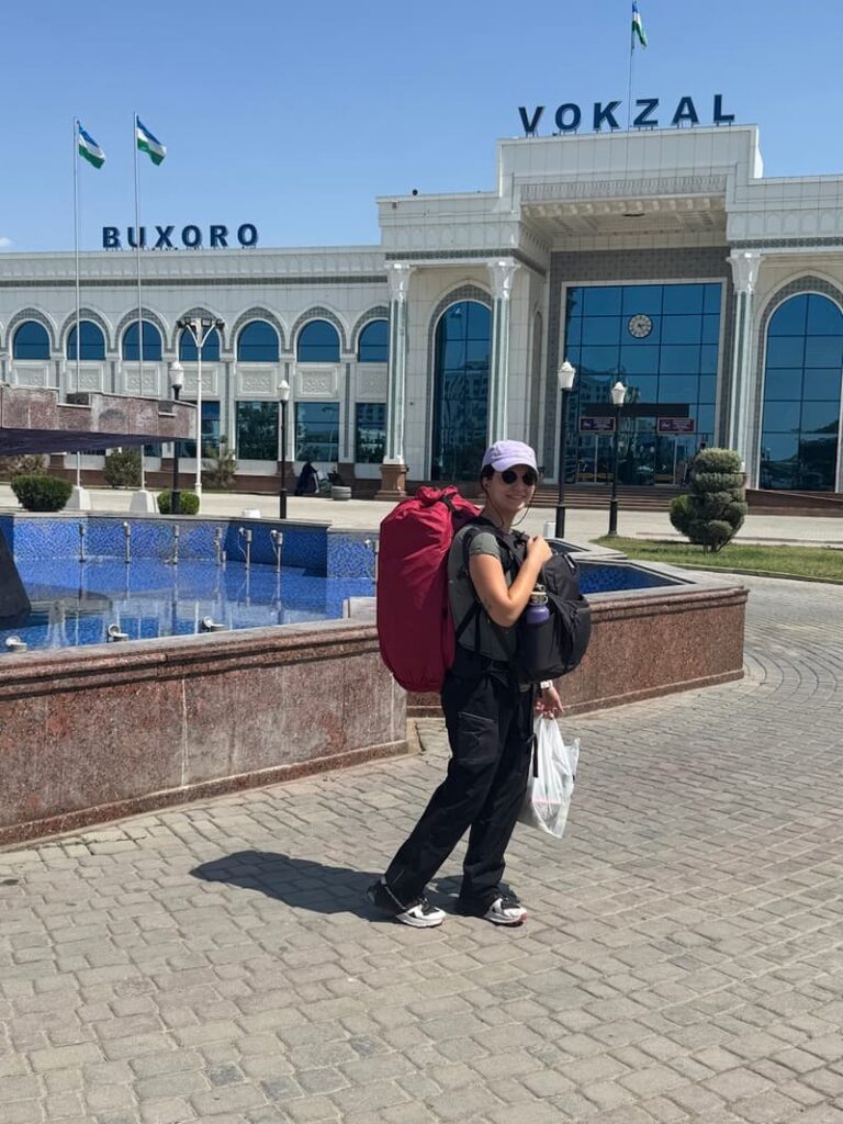 the author carrying a red backpack in the back and a black backpack in the front posing in front of the Bukhara train station in Uzbekistan