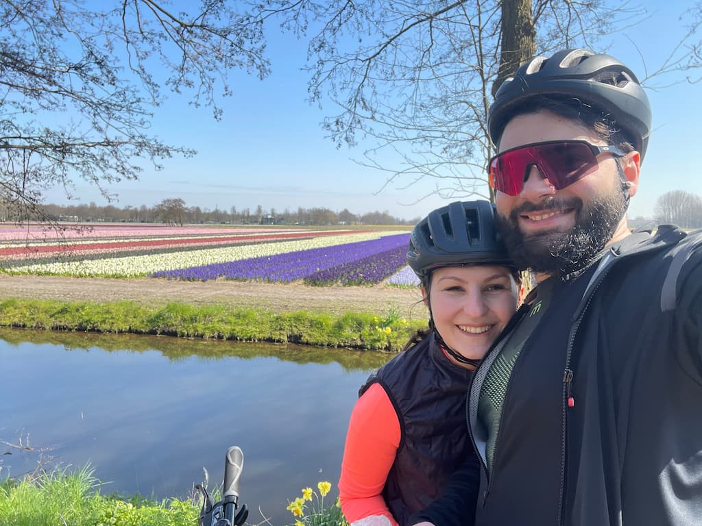 Eleni and Alessandro, the authors behind Beyond Our Footprints, wearing cycling clothing and equipment and posing in front of colourful tulip fields in the Netherlands, before quitting their jobs for long-term travel