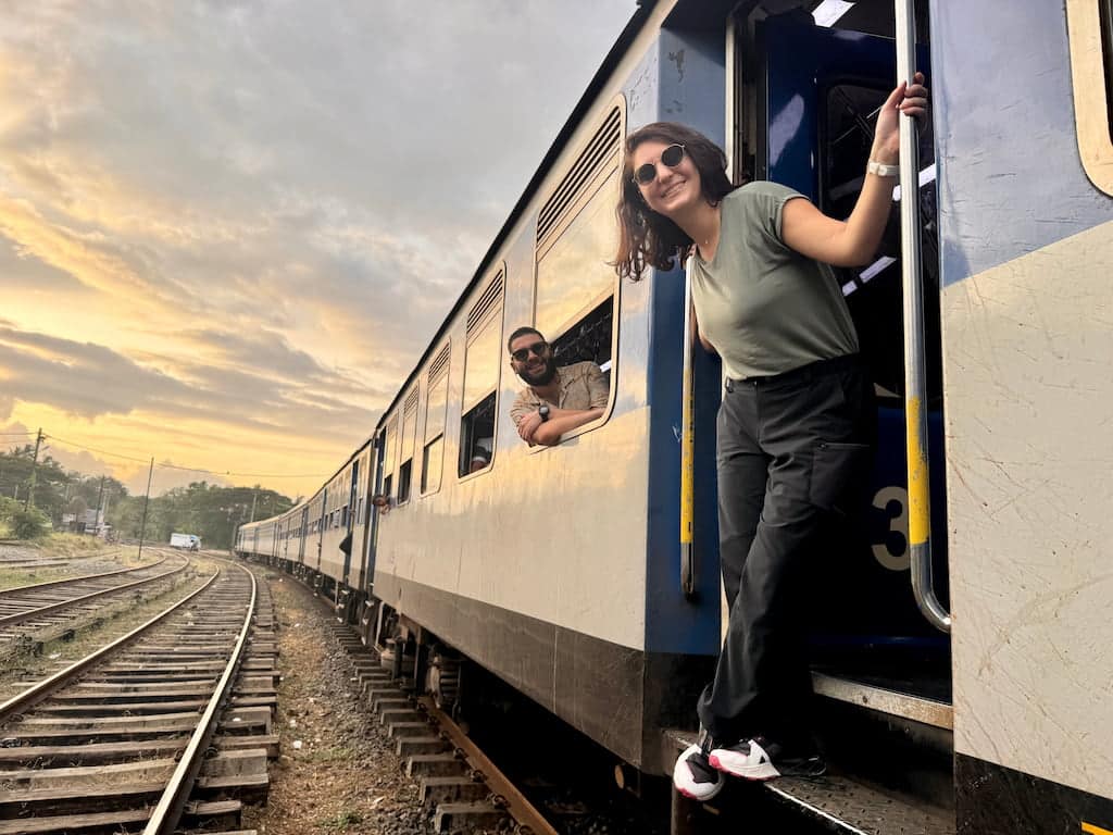 Eleni and Alessandro, the travel bloggers behind Beyond Our Footprints, hanging off a stopped train in Sri Lanka next to the rails
