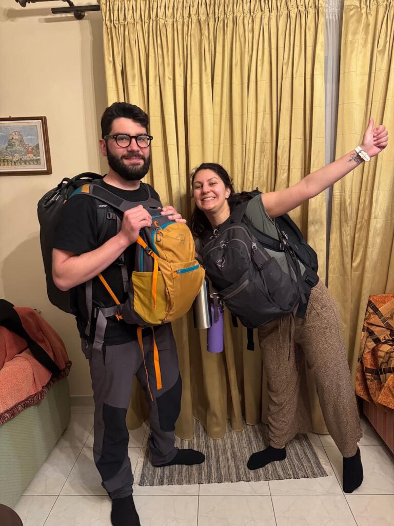 Eleni and Alessandro, travel bloggers behind Beyond Our Footprints, posing in a house holding all 4 backpacks and preparing to leave for their one-year trip