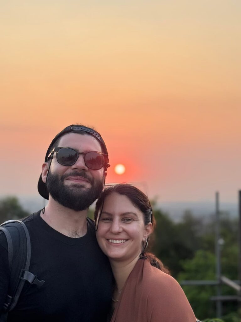 Portrait of Eleni and Alessandro, travel bloggers behind Beyond Our Footprints, smiling with a warm sunset in the back during their year-long world trip.