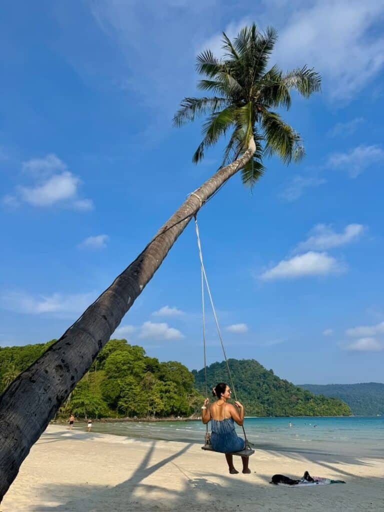 Eleni, author of the travel blog, swinging on a rope swing tied to a leaning coconut palm tree on a tropical white sand beach overlooking blue water and green hills.