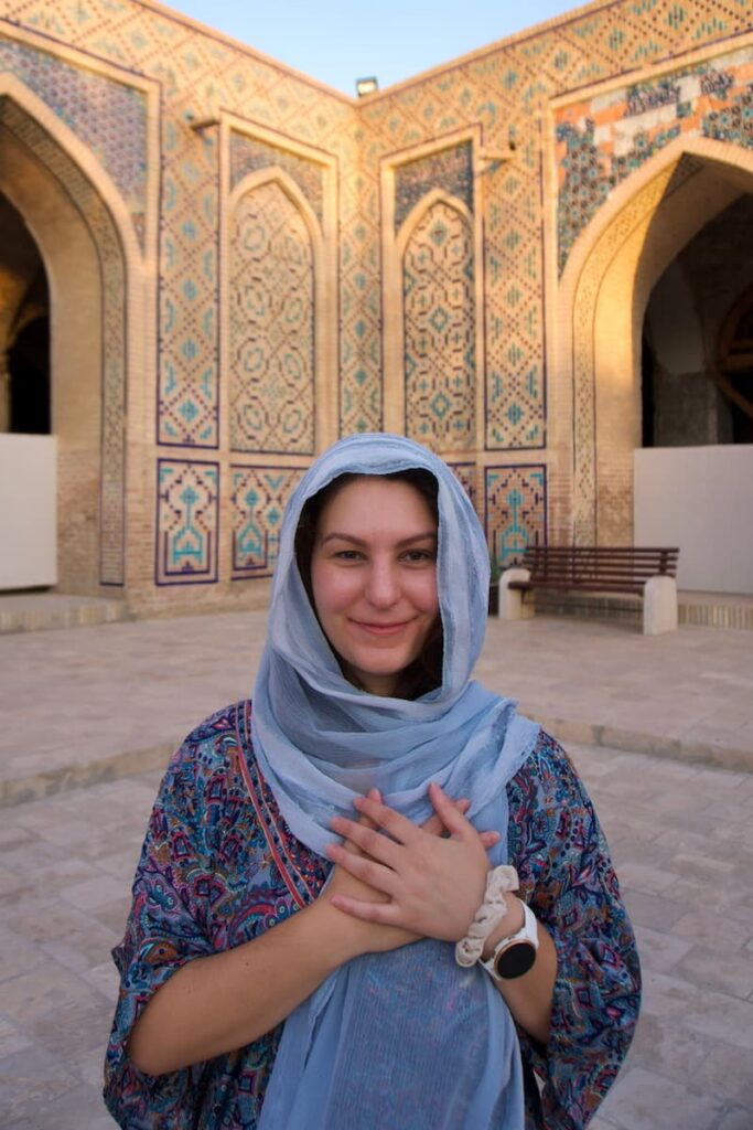Close-up portrait of a woman with her hands placed over her heart, standing before the intricate blue and tan geometric tile work of an ancient madrasa in Bukhara.