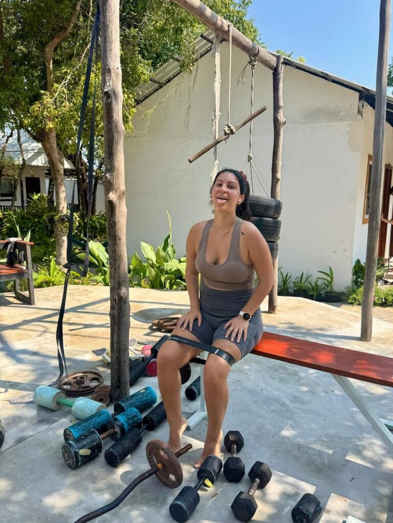 Eleni working out with a resistance band on a bench at an outdoor tropical resort gym surrounded by weights and equipment.
